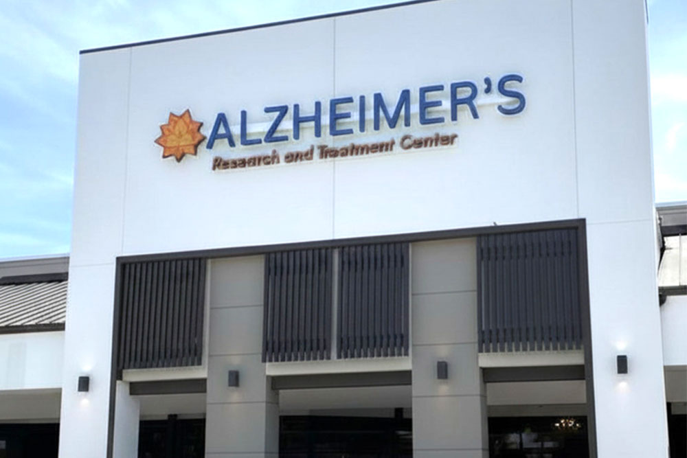 Front entrance of the Stuart Alzheimer's Research & Treatment Center, featuring a clear and welcoming sign with the center's name and address. The building is painted in light blue, with greenery in the foreground, representing a professional and inviting healthcare facility